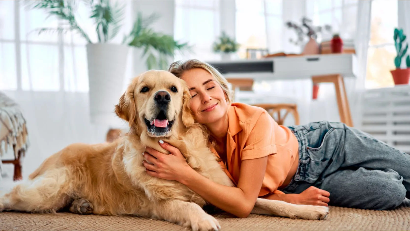 Woman smiling and hugging a Golden Retriever dog on the floor in a bright living room